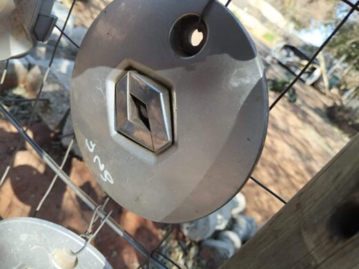 A close-up of a metallic hexagonal tool or part hanging on a wire fence, with a partially visible wooden post and a blurred backdrop of other auto parts and greenery.