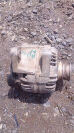 Front view of a used alternator for a small car engine, showing the pulley side and mounting points.Front view