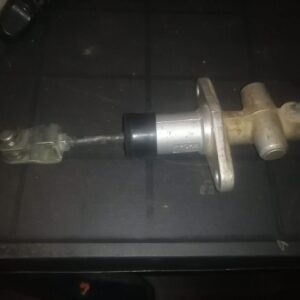 A metal automotive part, likely a clutch or brake master cylinder, featuring a cylindrical body, connecting rod, and attachment bracket, placed on a dark surface, possibly a workbench.
