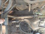 A close-up view of a vehicle's undercarriage showing various components such as pipes, a connector, and an actuator, with dirt and grass visible around the parts, indicating use or exposure to the outdoors.