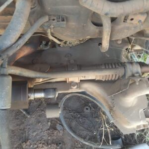 A close-up view of a vehicle's undercarriage showing various components such as pipes, a connector, and an actuator, with dirt and grass visible around the parts, indicating use or exposure to the outdoors.