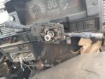 Close-up view of a car's steering column control, showing the steering wheel with gear and wiper control levers, part of a disassembled dashboard, and visible wiring beneath it.
