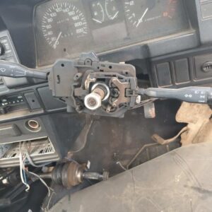 Close-up view of a car's steering column control, showing the steering wheel with gear and wiper control levers, part of a disassembled dashboard, and visible wiring beneath it.
