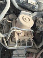 Close-up view of a dusty automotive part, likely a valve or actuator, with visible metal tubing and electrical connections, surrounded by cobwebs and dirt inside an engine compartment.
