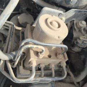 Close-up view of a dusty automotive part, likely a valve or actuator, with visible metal tubing and electrical connections, surrounded by cobwebs and dirt inside an engine compartment.