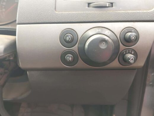 2012 Opel Corsa Essentia Headlight Switch – Used | Brits Brothers Control panel of a car interior featuring various switches including fog light controls, light dimmer, and a single large knob for headlight settings, with textured and matte surfaces.